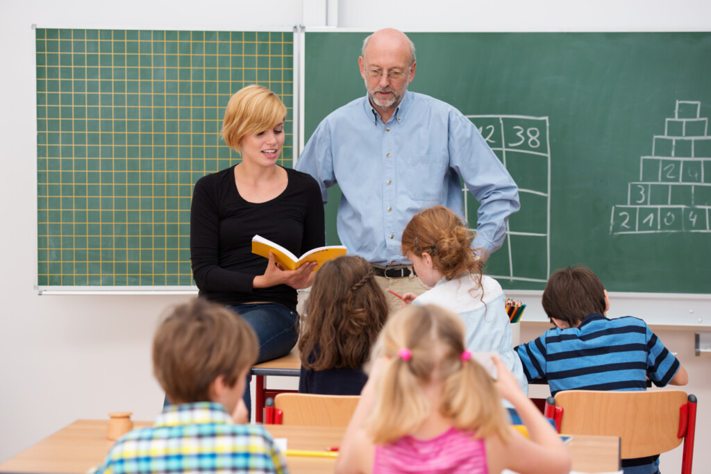 Teacher with his teaching assistant. Teacher with his young attractive female teaching assistant standing in front of a class of small girls and boys as they discuss something from a textbook